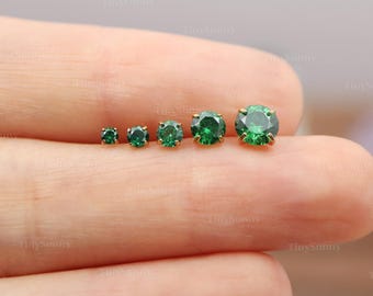 May include: Five round, brilliant-cut green gemstones set in gold-toned metal prongs, arranged in a row from smallest to largest. Each stone features a faceted surface that reflects light, showcasing a vibrant emerald green hue in a stud earring style.