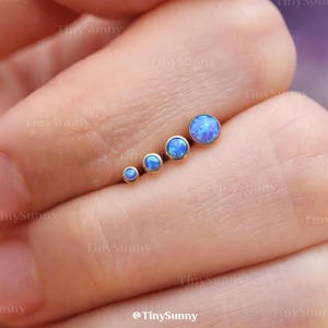 May include: A set of four graduated blue opal stud earrings. The earrings are arranged in a row, with the smallest on the left and the largest on the right. The earrings are set in a gold-colored metal.