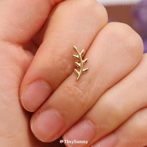 May include: A gold-colored, leaf-shaped earring stud is held between fingers. The earring has a delicate design with small leaves along a central stem. The background is a blurred view of a hand and a soft, neutral surface.