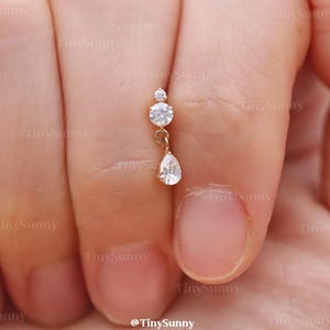 May include: A delicate gold earring featuring three clear, sparkling gemstones. The top has two round stones, and a pear-shaped stone dangles below. The earring is held up by a hand.