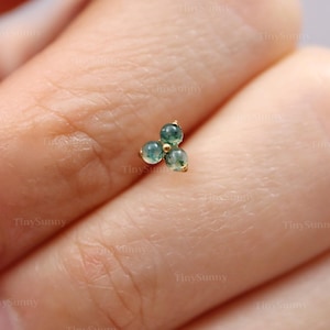 May include: A small, gold-colored stud earring featuring three round, teal-green gemstones arranged in a triangular cluster. The earring is set against a blurred background, highlighting the jewelry's delicate design.