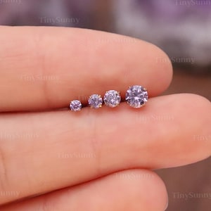 May include: A hand displays five small, round, light purple gemstones of varying sizes. The gemstones are set in a row, with the smallest on the left and the largest on the right. The background is blurred, with a hint of purple.