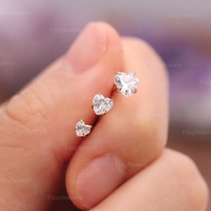 May include: Three gold-colored stud earrings, each featuring a clear, heart-shaped gemstone. The earrings are displayed in a hand, showcasing the varying sizes of the heart-shaped stones. The smallest earring is at the bottom, with the largest at the top.