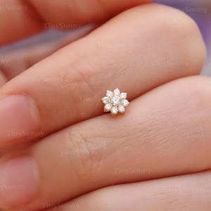 May include: A small, gold-colored flower-shaped stud earring with multiple clear gemstones. The earring is set against a skin-toned background. The text "@TinySunny" is visible.