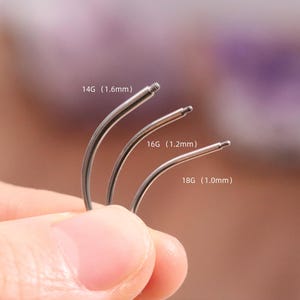 May include: Three curved, silver body jewellery pieces are shown, labelled with sizes 14G (1.6mm), 16G (1.2mm), and 18G (1.0mm). The jewellery is held by a hand against a blurred background.