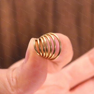 May include: A collection of gold-colored, spiral-shaped earrings. The earrings are held between fingers, showcasing their circular design and polished finish. The image is set against a soft, blurred background, highlighting the jewelry's details.