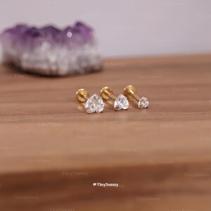 May include: Three gold-colored stud earrings with clear, heart-shaped cubic zirconia stones. The earrings are displayed on a wooden surface. The smallest earring is a single clear stone. The brand name "TinySunny" is visible.