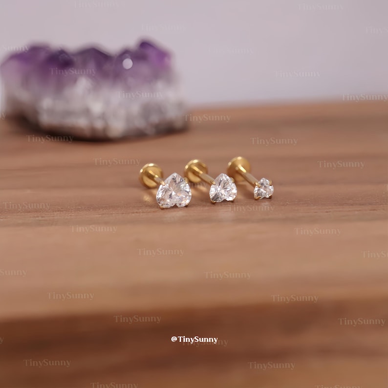 May include: Three gold-colored stud earrings with clear, heart-shaped cubic zirconia stones. The earrings are displayed on a wooden surface. The smallest earring is a single clear stone. The brand name "TinySunny" is visible.