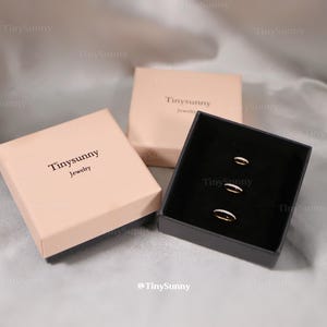 May include: Three rings with small clear stones are displayed in a black box. Two closed pink boxes with the text "Tinysunny Jewelry" are also in the image. The background is a light gray fabric.