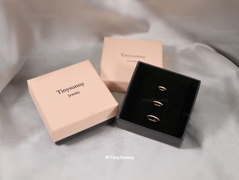 May include: Three rings with small clear stones are displayed in a black box. Two closed pink boxes with the text "Tinysunny Jewelry" are also in the image. The background is a light gray fabric.