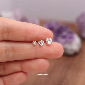 May include: Three heart-shaped stud earrings in varying sizes. The earrings are clear and sparkly, set in gold-colored metal. The earrings are displayed on a light-colored background.