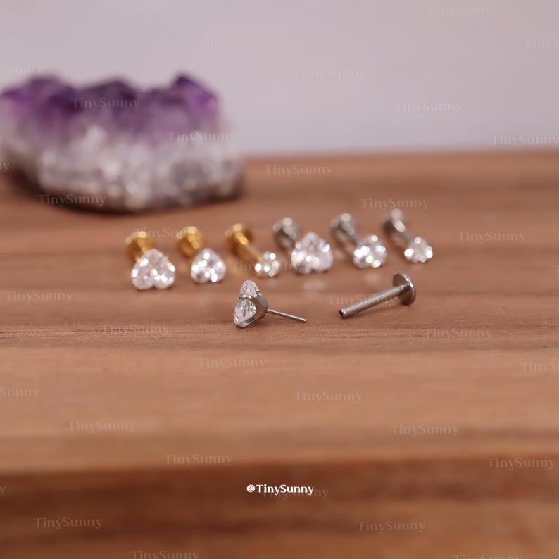 May include: A collection of heart-shaped stud earrings and a single labret stud with a heart-shaped gem. The earrings are in gold and silver tones, with clear, sparkling gems. The background is a wooden surface.