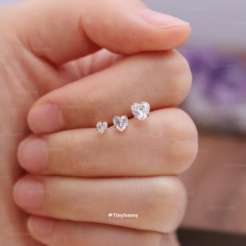 May include: Three heart-shaped stud earrings with clear, sparkling stones. The earrings are displayed on a hand, showcasing their varying sizes. The smallest earring is on the left, followed by a medium-sized one, and a larger one on the right. The earrings are likely made of silver or white gold.