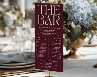 May include: A vertical rectangular menu sign with a deep burgundy background and white text reading THE BAR, Signature Cocktails, The Classics, Wine & Beer, and Non Alcoholic. The sign is mounted on a thin gold metal stand with a square base.