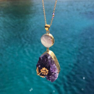 Puede incluir: Un collar de cadena dorada con un colgante que presenta un gran cristal de amatista y una perla blanca redonda m&aacute;s peque&ntilde;a. El cristal de amatista es un c&uacute;mulo de cristales morados con detalles dorados.