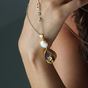 Puede incluir: Un collar de cadena dorada con un colgante que presenta una perla blanca y una piedra p&uacute;rpura con un dise&ntilde;o de flor dorada.
