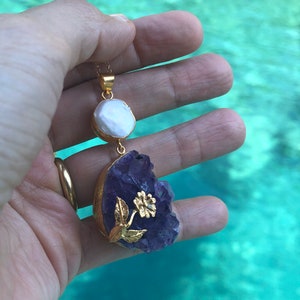 Puede incluir: Un collar de oro con un colgante de cristal de amatista p&uacute;rpura y un dije de perla blanca. El colgante presenta un dise&ntilde;o de flor de oro.