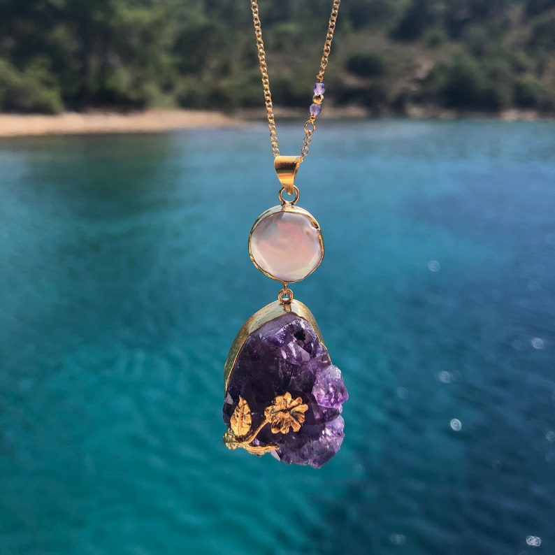 Puede incluir: Un collar de oro con una cadena delicada y un colgante que presenta un gran cristal de amatista con detalles dorados y una piedra redonda m&aacute;s peque&ntilde;a de color rosa p&aacute;lido.