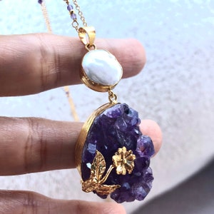 Puede incluir: Un collar de oro con un gran colgante de cristal de amatista y un peque&ntilde;o colgante de perla. El cristal de amatista est&aacute; rodeado de hojas de oro y una flor.
