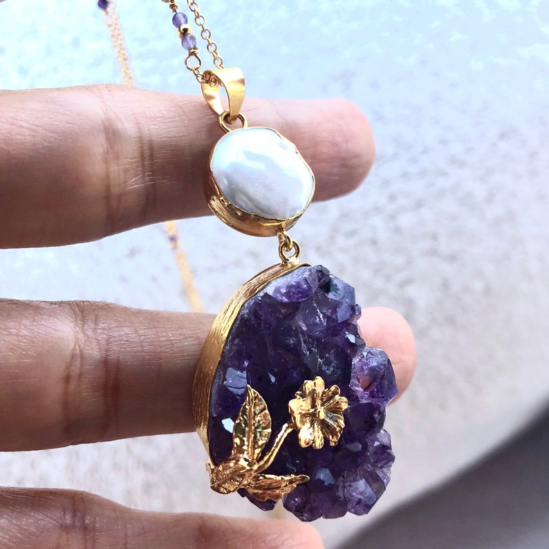 Puede incluir: Un collar de oro con un gran colgante de cristal de amatista y un peque&ntilde;o colgante de perla. El cristal de amatista est&aacute; rodeado de hojas de oro y una flor.
