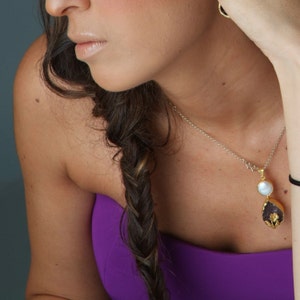 Puede incluir: Una mujer con el pelo largo y casta&ntilde;o trenzado de un lado, lleva un top morado sin tirantes y un collar de cadena plateada con un colgante de oro con una perla blanca y una piedra morada.
