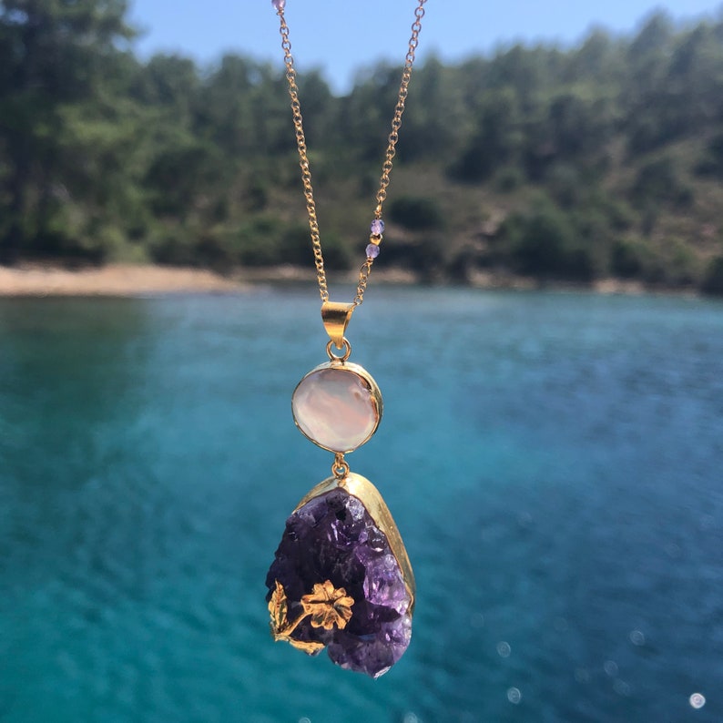 Puede incluir: Un collar de cadena dorada con un gran colgante de cristal de amatista p&uacute;rpura y un colgante m&aacute;s peque&ntilde;o de n&aacute;car blanco. El cristal de amatista es en bruto y tiene una forma natural irregular. El collar est&aacute; fotografiado sobre un fondo de agua azul y &aacute;rboles verdes.