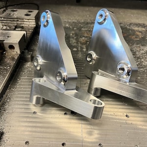 May include: Two machined aluminum brackets with multiple holes and a silver finish. The brackets have a triangular shape with a rectangular base and are designed for mechanical applications. The image shows the parts on a metal surface with machining marks.