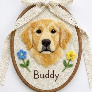 以下が含まれることがあります： ゴールデンレトリバーの肖像画が描かれた、手作りの楕円形の壁掛け。犬の名前「Buddy」が下に刺繍されています。木製のフープに額装され、レースのリボンと2つの小さなカラフルな花のアクセントで飾られています。