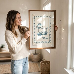 May include: A framed artwork featuring a dachshund wearing a cap and riding a skateboard, with the name "Winnie" below. The artwork is in a wooden frame with a cream-colored background and blue embroidered details. The frame is being held up.