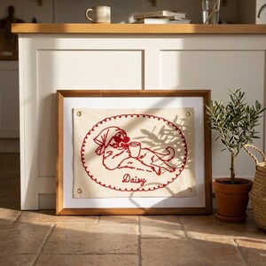 May include: A framed embroidered artwork featuring a red cat design. The cat, wearing a headscarf and sunglasses, is holding a mug. The word "Daisy" is embroidered below the cat. The frame is wood.
