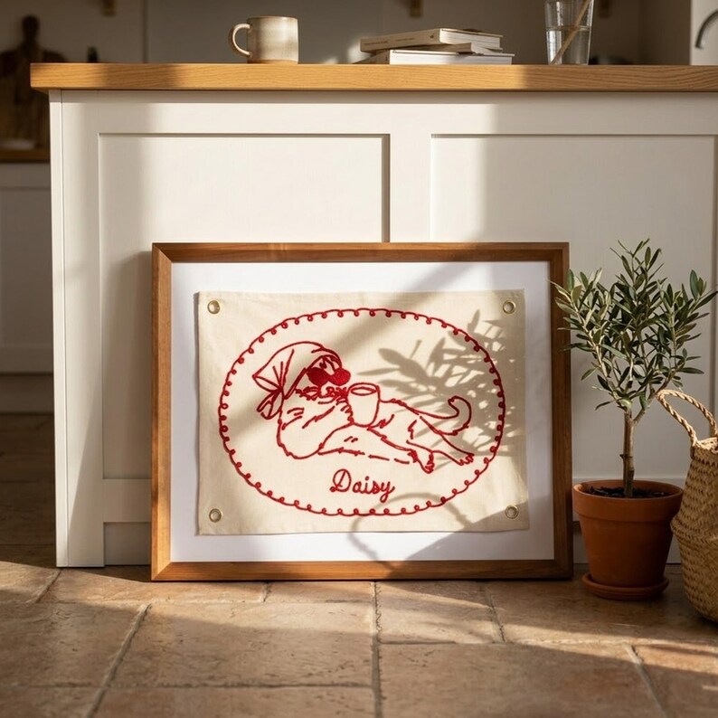 May include: A framed embroidered artwork featuring a red cat design. The cat, wearing a headscarf and sunglasses, is holding a mug. The word "Daisy" is embroidered below the cat. The frame is wood.