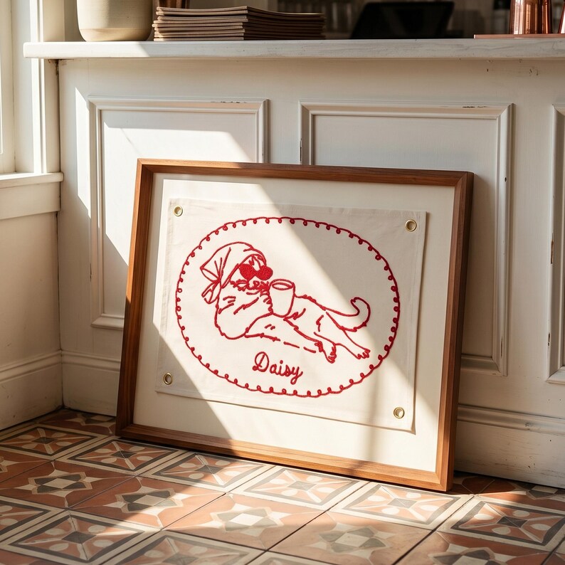 May include: A framed artwork featuring a red embroidered illustration of a cat wearing sunglasses and a headscarf, holding a cup. The cat is inside an oval with a scalloped border, with the name "Daisy" below. The frame is made of wood.