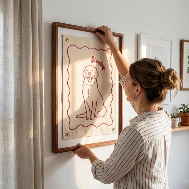 以下が含まれることがあります： 犬のイラストと「Simba」という名前が赤で刺繍された、額入りのキャンバスウォールハンギング。アートワークは濃い木製のフレームにセットされ、白い壁に掛けられています。