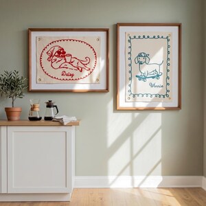 May include: Two framed dog art prints. One features a red embroidered dog with the name "Daisy". The other shows a blue embroidered dog on a skateboard with the name "Winnie". Both are in wooden frames.