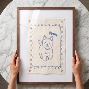 以下が含まれることがあります： 犬のイラストが描かれた額入りのアートワーク。アートワークは、青い刺繍の犬のデザインと、その上に「Nala」という名前が書かれた、明るいベージュ色の生地です。フレームは濃い茶色の木で、アートワークは白いマットに展示されています。