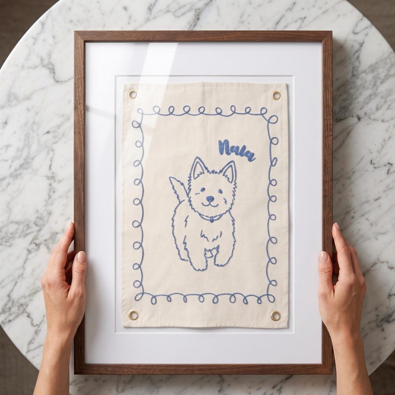 以下が含まれることがあります： 犬のイラストが描かれた額入りのアートワーク。アートワークは、青い刺繍の犬のデザインと、その上に「Nala」という名前が書かれた、明るいベージュ色の生地です。フレームは濃い茶色の木で、アートワークは白いマットに展示されています。