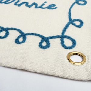 以下が含まれることがあります： クリーム色の生地のバナーで、青い糸で「Winnie」と刺繍されています。バナーには装飾的な青い刺繍の縁取りと金のグロメットが付いています。