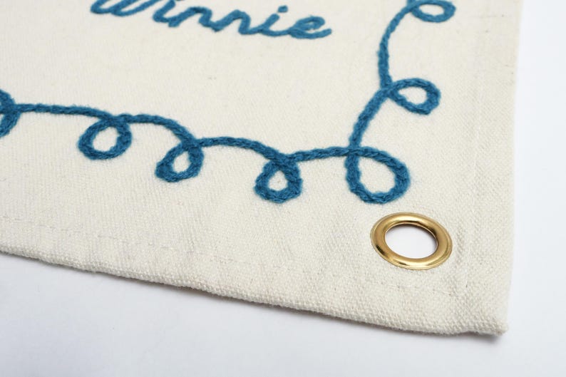 以下が含まれることがあります： クリーム色の生地のバナーで、青い糸で「Winnie」と刺繍されています。バナーには装飾的な青い刺繍の縁取りと金のグロメットが付いています。