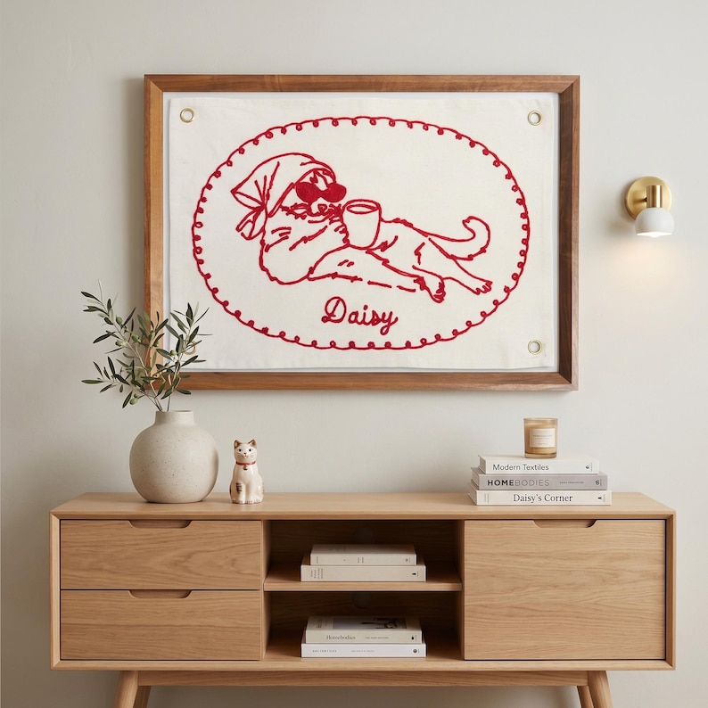 May include: A framed wall hanging featuring a red embroidered design of a dog wearing sunglasses and a headscarf, holding a cup. The dog's name, "Daisy," is embroidered below. The frame is wood, and the background is white.