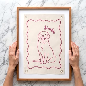 以下が含まれることがあります： 犬のイラストと「Simba」の名前が赤紫で刺繍された、額装されたキャンバスアート。犬とボーダーは同じ色です。フレームは木製です。