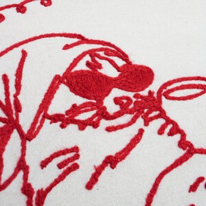 May include: Close-up of a white fabric with a red embroidered design. The embroidery depicts a whimsical, cartoon-like figure with a hat and a cup. The stitching is thick and textured, creating a bold visual effect.