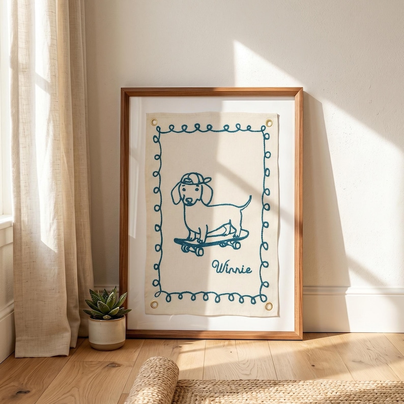 May include: A framed artwork featuring a dachshund dog on a skateboard, embroidered in teal thread on a cream-colored background. The dog wears a cap and the name "Winnie" is written below. The frame is made of wood.