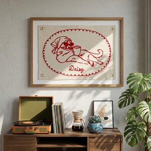 May include: A framed wall hanging with a red embroidered design of a dog wearing a cap and sunglasses, holding a mug, with the name "Daisy" below. The frame is wood-toned. A record player, vinyl records, and a plant are also visible.