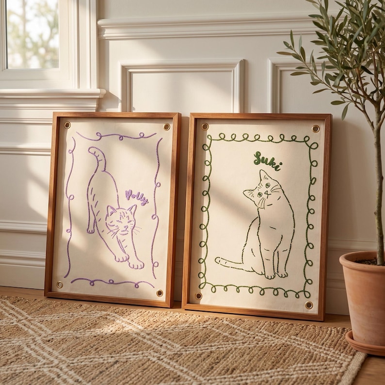 以下が含まれることがあります： 2つの額入り猫アート作品。1つは「Polly」という名前の紫色の刺繍猫が特徴です。もう1つは「Suki」という名前の緑色の刺繍猫が特徴です。どちらも木製のフレームが付いたクリーム色の背景にあります。