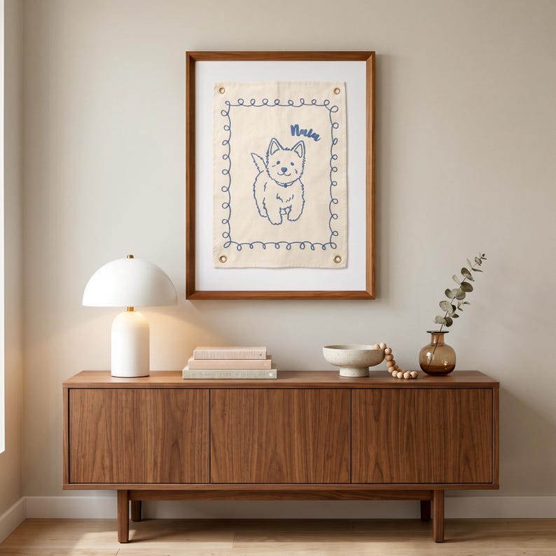 以下が含まれることがあります： クリーム色の背景に、青い刺繍で犬のイラストと「Nala」という名前が描かれた額入りのアートワーク。白いランプ、本、ボウル、花瓶のある木製のキャビネットの上に飾られています。