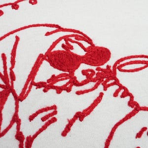 以下が含まれることがあります： 白い生地に赤い刺繍が施されたクローズアップ。刺繍は、帽子とカップのある顔の様式化されたデザインを示しています。デザインは、太く、質感のある赤い糸で作られています。