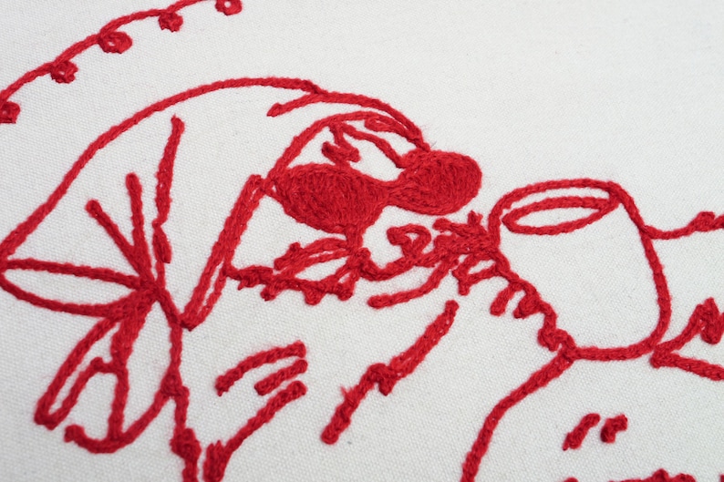 以下が含まれることがあります： 白い生地に赤い刺繍が施されたクローズアップ。刺繍は、帽子とカップのある顔の様式化されたデザインを示しています。デザインは、太く、質感のある赤い糸で作られています。