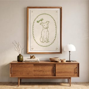 以下が含まれることがあります： ベンジという名前の犬の線画が描かれた額入りのアートワーク。犬は装飾的なボーダーが付いた緑色の楕円の中にいます。アートワークは明るい色の生地に取り付けられ、木で額装されています。アートワークは木製のキャビネットの上に掛けられています。