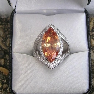 May include: A silver ring with a large, marquise-cut, orange gemstone. The gemstone is surrounded by a halo of smaller, clear stones. The ring is set in a silver band and is displayed in a black jewelry box with a white satin interior.