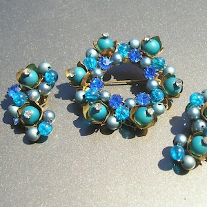 May include: A set of vintage jewelry featuring a large turquoise and blue brooch with a circular design and matching earrings. The brooch is made of gold-toned metal and features a variety of turquoise and blue beads and rhinestones.